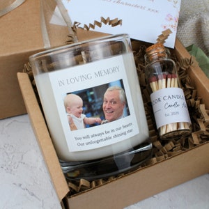 May include: A white candle in a glass jar with a photo of a man and a child. The candle has the text "In Loving Memory" and "You will always be in our hearts. Our unforgettable shining star." The candle is in a brown box with shredded paper. There is a small glass jar with matches that says "Cor Candle - Candle A Est. 2020".