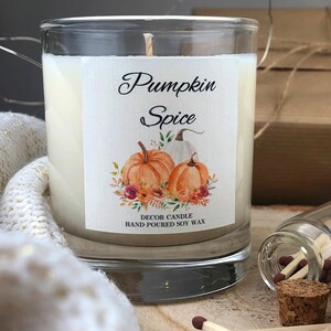 May include: A glass jar candle with a white label that says "Pumpkin Spice" and features a watercolor illustration of pumpkins and fall foliage. The label also says "Decor Candle Hand Poured Soy Wax".