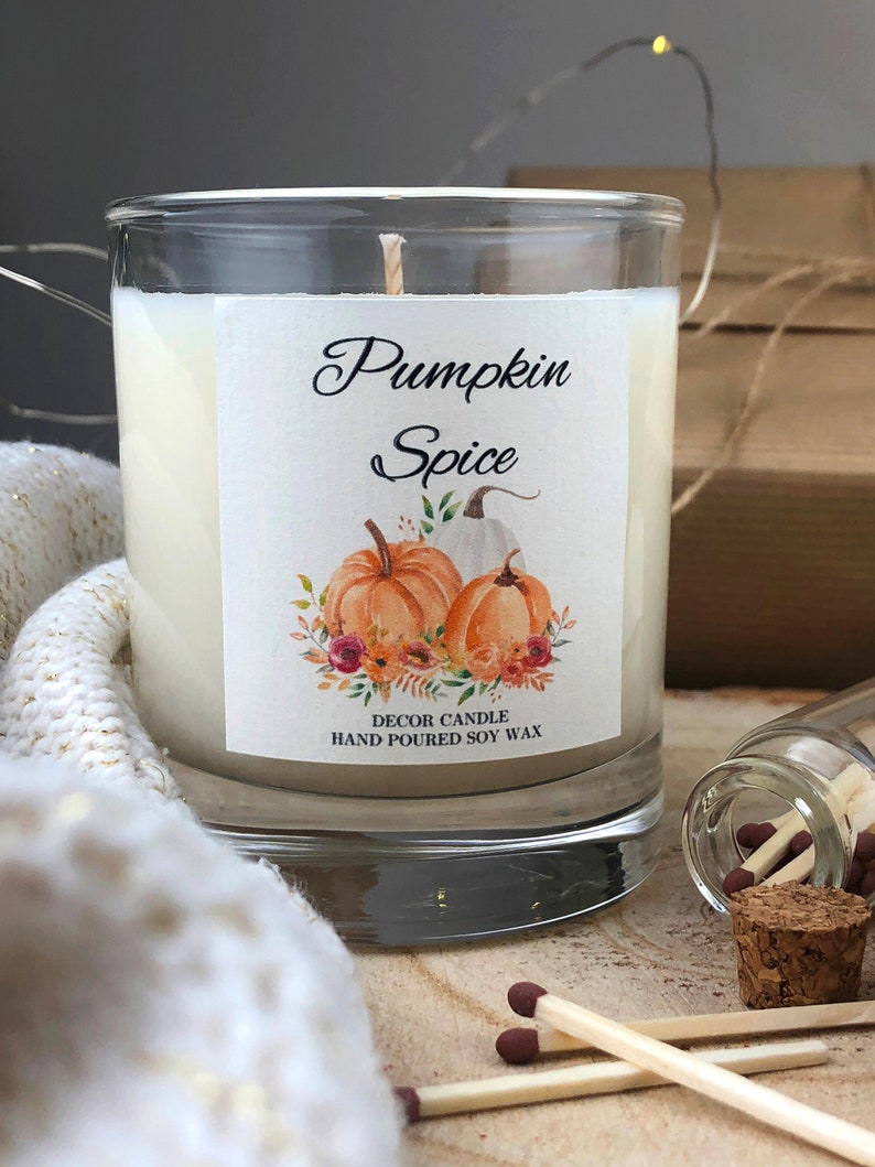 May include: A glass jar candle with a white label that says "Pumpkin Spice" and features a watercolor illustration of pumpkins and fall foliage. The label also says "Decor Candle Hand Poured Soy Wax".