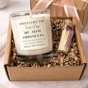 May include: A clear glass candle with a white label reading "SMELLS LIKE THE Tears of my JIU-JITSU OPPONENTS infused with a hint of victory and pure champion confidence Happy 30th Andrew!" next to a small glass bottle of matches in a brown box.