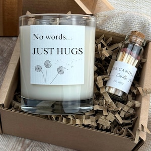 May include: A candle in a clear glass jar with the text "No words... JUST HUGS" on a white label. The candle sits in a brown cardboard box with shredded paper. A small glass bottle of matches is also in the box.