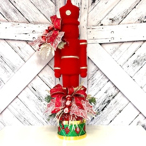 May include: A red velvet nutcracker with gold trim stands on a green and gold base with a red and white bow and greenery.