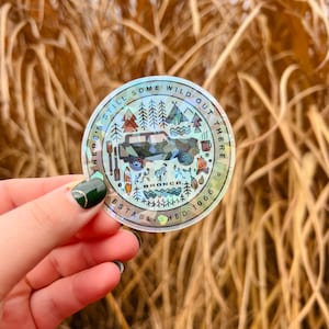 May include: A round, holographic sticker with the text "THERE'S STILL SOME WILD OUT THERE" and "ESTABLISHED 1966". The sticker features a graphic of a camouflage-colored vehicle, trees, mountains, and camping elements. The sticker is held by a hand.