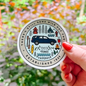 Może przedstawiać: Okrągła naklejka z niebieskim Toyota 4Runner, drzewami, namiotem, ogniskiem i kajakiem. Naklejka zawiera napis "There's still some wild out there" i "Established 1984".