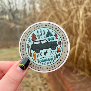 Peut inclure: Un autocollant rond avec un Ford Bronco vert conduisant à travers une scène de forêt. L'autocollant dit "There's still some wild out there. Established 1966. Bronco."