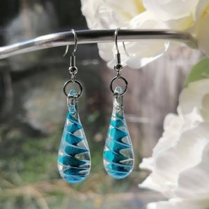 Puede incluir: Un par de pendientes de vidrio en forma de lágrima con un diseño de espiral azul y blanco. Los pendientes son plateados y tienen un cierre de gancho.