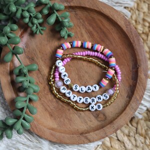 May include: A set of three stacked beaded bracelets. One bracelet features multi-coloured striped clay beads, another has solid pink beads, and the third consists of gold-toned beads. White letter beads spell out the words LIKE SLAY PATTERNS KB.