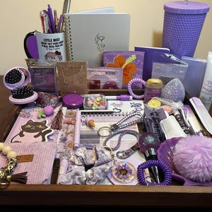 Peut inclure: Une collection d'accessoires violets, dont un gobelet, une tasse, un carnet, des bijoux, une brosse à cheveux et des chouchous, disposés sur un plateau en bois. Les articles sont de différentes nuances de violet, créant une esthétique cohérente.