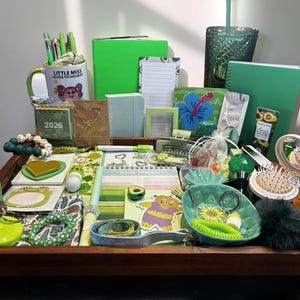 Peut inclure: Une collection de papeterie et d'accessoires sur le thème du vert, comprenant des cahiers, des stylos, une tasse et un agenda quotidien. Les articles sont disposés dans un plateau en bois, avec une variété de nuances de vert et quelques accents d'autres couleurs.