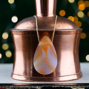 Peut inclure: Un pendentif en forme de goutte avec des motifs tourbillonnants orange et blancs est suspendu à une fine chaîne dorée. Le pendentif repose sur une bouteille en cuivre poli avec un bouchon en liège, dotée d'une finition métallique lisse.
