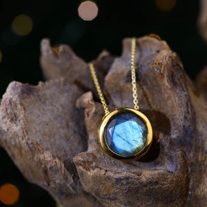 May include: A gold pendant necklace featuring a round, faceted labradorite gemstone with blue iridescence. The stone is set in a polished gold bezel and hangs from a delicate gold chain, showcasing a smooth, reflective surface and deep blue hues.