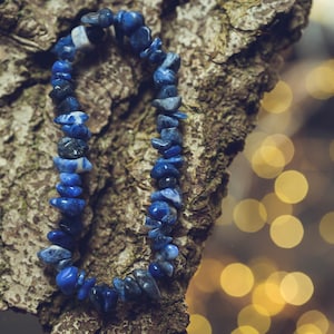 Könnte beinhalten: Ein Armband bestehend aus zahlreichen unregelmäßigen, getrommelten blauen Steinperlen. Die Perlen weisen verschiedene Schattierungen von tiefem Blau, Marineblau und gelegentlichen weißen Marmorierungen auf, die zu einer Schlaufe gereiht sind.