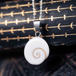 Op de afbeelding: Een zilverkleurige ketting met een gladde, witte, ronde hanger met een natuurlijk bruin spiraalpatroon. De hanger is met een eenvoudig metalen oogje aan de ketting bevestigd, wat zorgt voor een strakke en gebalanceerde uitstraling van dit sieraad.