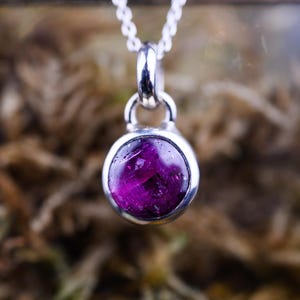 Op de afbeelding: Een ronde, diep magenta edelsteen hanger gezet in een gepolijste zilveren zetting. De steen heeft een natuurlijk getextureerd oppervlak en hangt aan een fijne zilveren ketting via een eenvoudig, afgerond zilveren oogje voor een strak sieraad.