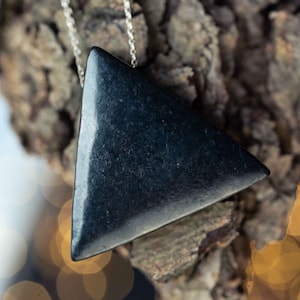 Pode incluir: Um colar com um pendente triangular liso de cor cinzento-carvão escuro, suspenso por uma fina corrente de metal prateado. O pendente tem uma superfície mate ligeiramente texturada e a forma de um triângulo equilátero com cantos arredondados.