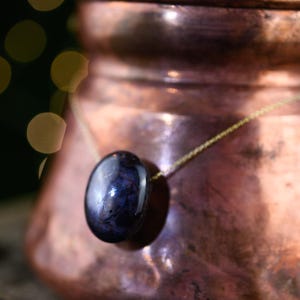 Könnte beinhalten: Eine Halskette mit einem glatten, oval geformten, dunkelvioletten Edelsteinanhänger, der an einer dünnen, zierlichen goldfarbenen Metallkette hängt. Der Stein hat eine tiefe, reflektierende Oberfläche mit subtilen Farbnuancen.