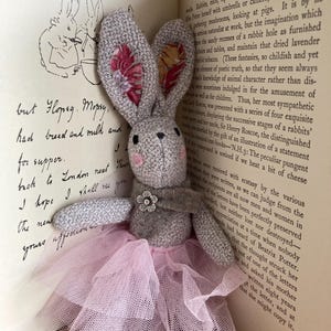 May include: A handcrafted grey bunny toy with a pink tulle skirt and a flower detail. The bunny's ears have a floral pattern. It is placed beside an open book with text and illustrations, reminiscent of a children's story.