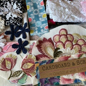 May include: A selection of fabric pieces and decorative elements, featuring embroidered floral patches, lace, and patterned textiles. The colour scheme includes white, black, blue, pink, and burgundy. A tag reads "Cardboard & Scraps by Crafting with Lottie."