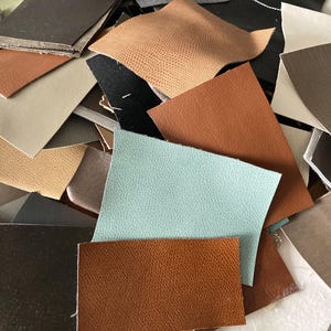 May include: A pile of leather offcuts in a variety of colours and textures. The pieces include shades of brown, tan, black, and a light blue. Some pieces have a textured surface, while others are smooth. The offcuts are of different shapes and sizes.