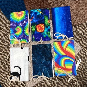 May include: Six fabric-covered journals with various designs. The covers feature a tie-dye pattern, a space scene with rockets and planets, a blue galaxy, a black cat silhouette, a dark blue floral pattern, and a rainbow design. Each journal has a string closure.