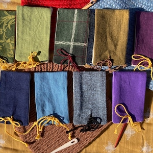 May include: A collection of handmade fabric book covers in various colors and patterns. The covers include green floral, solid green, plaid, tan, purple, navy, light blue, gray, and purple. Each cover is tied with a coordinating cord.