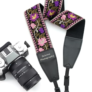 May include: A black camera strap with embroidered floral design in pink, purple, and gold. The strap has black leather ends and the text "Sabrina Photography". A silver camera with a black lens is visible.