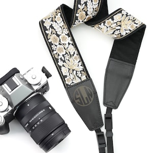 Può includere: Tracolla per fotocamera nera con ricamo floreale dorato e bianco. La tracolla ha una sezione in pelle nera con un monogramma "SLN" in oro.
