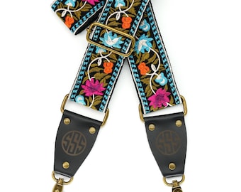 May include: A black camera strap with a floral pattern in shades of pink, blue, orange, and green. The strap has two leather ends with brass buckles and a monogrammed 'SSS' on each end.
