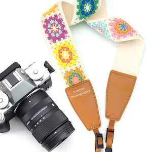 May include: A white camera strap with colourful crocheted squares and a brown leather pad with the text "Sabrina Photography".