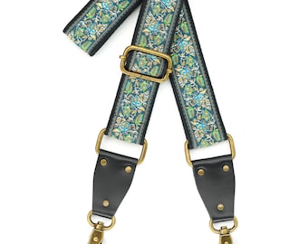 May include: A teal and gold patterned camera strap with black leather accents and brass hardware. The strap has two swivel hooks.