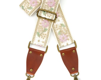 May include: A white camera strap with pink and green floral embroidery, brown leather ends, and gold metal hardware.
