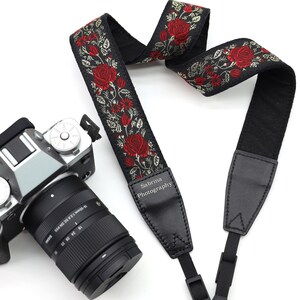 May include: Black camera strap with a red and gold floral pattern. The strap has a black leather patch with the text "Sabrina Photography" on it.