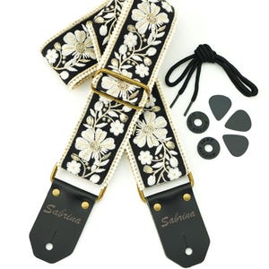 Puede incluir: Correa de guitarra negra con bordado floral blanco y herrajes dorados. La correa tiene extremos de cuero negro con el nombre "Sabrina" impreso. Incluye púas negras, cierres de correa y un cordón negro.