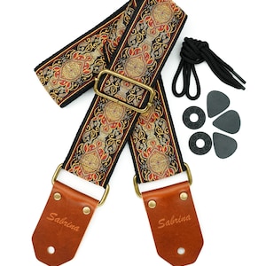 Puede incluir: Una correa de guitarra negra con un patrón bordado en oro y rojo. La correa tiene extremos de cuero marrón con la palabra "Sabrina" grabada en relieve. La correa viene con una púa de guitarra negra y un cierre de correa negro.