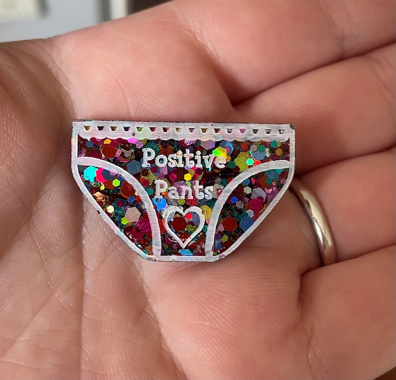 Pu&ograve; includere: Uno spillo in resina glitterata colorata a forma di biancheria intima con il testo "Positive Pants" e un piccolo cuore al centro.