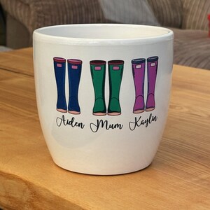 May include: White ceramic planter featuring a design of three pairs of colourful rain boots. The boots are labelled "Aiden", "Mum", and "Kaylin".