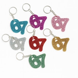 May include: A selection of glittery keyrings, each displaying the number '67'. The keyrings come in a range of colours: teal, purple, pink, silver, red, gold, and green. Each has a silver-coloured chain and key ring.