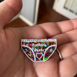 Pu&ograve; includere: Uno spillo in resina bianca riempito di glitter a forma di biancheria intima con il testo "Positive Pants" e un piccolo cuore al centro.