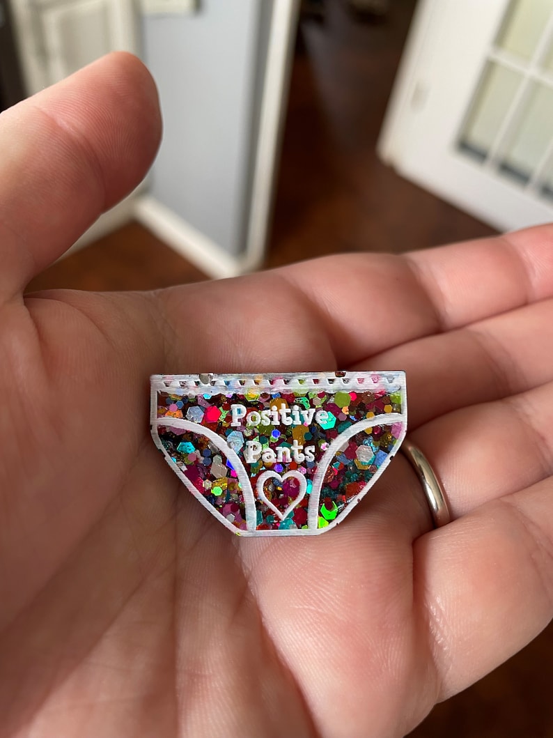 Pu&ograve; includere: Uno spillo in resina bianca riempito di glitter a forma di biancheria intima con il testo "Positive Pants" e un piccolo cuore al centro.