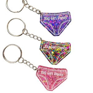 Peut inclure: Trois porte-cl&eacute;s avec des breloques en r&eacute;sine remplies de paillettes en forme de sous-v&ecirc;tements. Chaque breloque porte l'inscription "Big Girl Pants". Les breloques sont violettes, multicolores et roses.