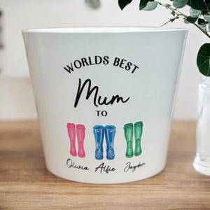 May include: White ceramic planter with the text "Worlds Best Mum To" and three pairs of colourful wellies with names Olivia, Alfie, and Jayden written below.
