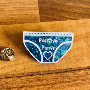 Pu&ograve; includere: Uno spillo di glitter blu a forma di biancheria intima con il testo "Positive Pants" e un cuore bianco al centro.