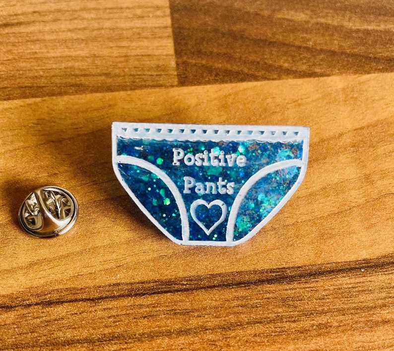 Pu&ograve; includere: Uno spillo di glitter blu a forma di biancheria intima con il testo "Positive Pants" e un cuore bianco al centro.
