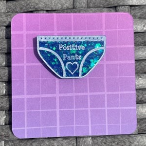 Pu&ograve; includere: Uno spillo in resina blu con glitter a forma di biancheria intima con il testo "Positive Pants" e un cuore bianco al centro.