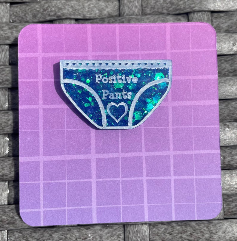 Pu&ograve; includere: Uno spillo in resina blu con glitter a forma di biancheria intima con il testo "Positive Pants" e un cuore bianco al centro.