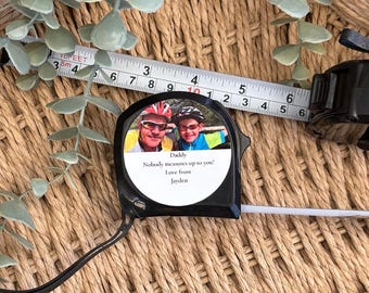May include: A black tape measure with a personalized label featuring a photo of two people wearing bike helmets and the text "Daddy Nobody measures up to you! Love from Jayden".