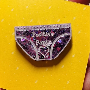 Pu&ograve; includere: Uno spillo in resina con glitter viola a forma di biancheria intima con il testo "Positive Pants" e un piccolo cuore al centro.