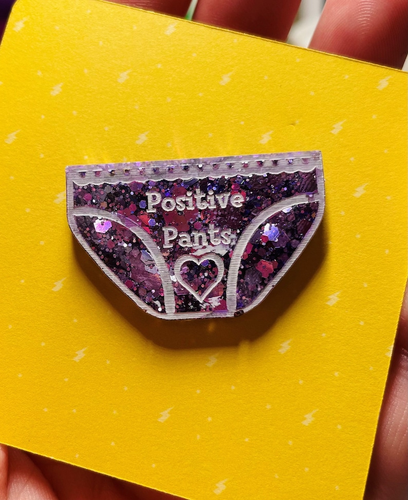 Pu&ograve; includere: Uno spillo in resina con glitter viola a forma di biancheria intima con il testo "Positive Pants" e un piccolo cuore al centro.