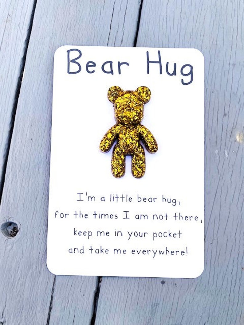 May include: A white card with the text "Bear Hug" in black lettering. A small, gold glitter bear is glued to the card. The text below the bear reads "I'm a little bear hug, for the times I am not there, keep me in your pocket and take me everywhere!"