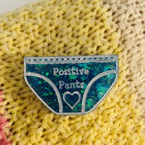 Pu&ograve; includere: Uno spillo di glitter blu e verde a forma di biancheria intima con il testo "Positive Pants" e un cuore al centro.